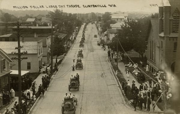 Elevated view of a Labor Day parade with more than thirty identical automobiles in a long line. Crowds are watching from the sidewalk. Caption reads: "Million Dollar Labor Day Parade, Clintonville, Wis."