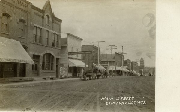 View looking across unpaved Main Street towards commercial buildings, including a meat market and the First National Bank. There are horse-drawn vehicles parked along the curb. Caption reads: "Main Street, Clintonville, Wis."