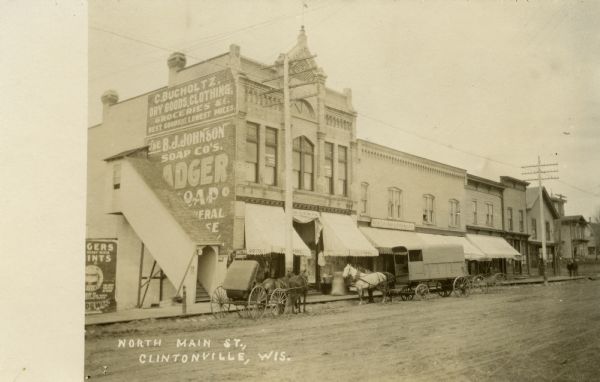 Black and white photograph looking across unpaved North Main Street towards commercial buildings. Two horse-drawn carriages and one horse-drawn freight wagon are parked along the curb. Signs are painted on the sides of the buildings, with one sign reading: "C. Bucholtz, Dry Goods, Clothing & Groceries, Best Goods At Lowest Prices." Another sign reads: "B.J. Johnson Soap Co's." Caption reads: "North Main St., Clintonville, Wis."