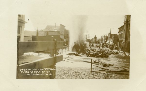 View down street towards the dynamiting of the bridge over the Pigeon River during the flood. Caption reads: "Dynamiting the Bridge, Flood of July 24, 1912, Clintonville, Wis."