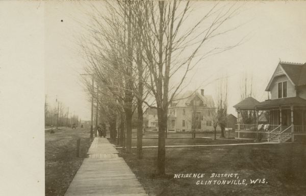 View down wooden sidewalk towards an unpaved street on the left, and a residential are with houses with large porches on the right. A group of people are walking up the sidewalk, and there are wagons in the street. Caption reads: "Residence District, Clintonville, Wis."