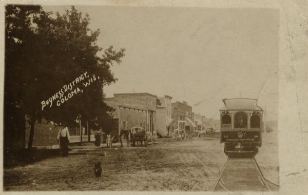 Photographic postcard of the business district. On the right is a streetcar, in the center is a horse and buggy, along with pedestrians and several commercial buildings. Caption reads: "Business District, Coloma, Wis."