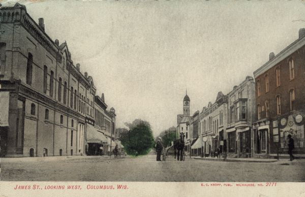 View down center of street looking west along James Street. There are pedestrians and two horse-drawn carriages on the street. Caption reads: "James St., Looking West, Columbus, Wis."