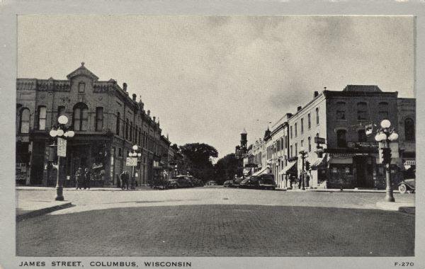 Black and white photograph looking west down James Street at intersection, with pedestrians on the sidewalks and parked automobiles along the curbs. Caption reads: "James Street, Columbus, Wis."