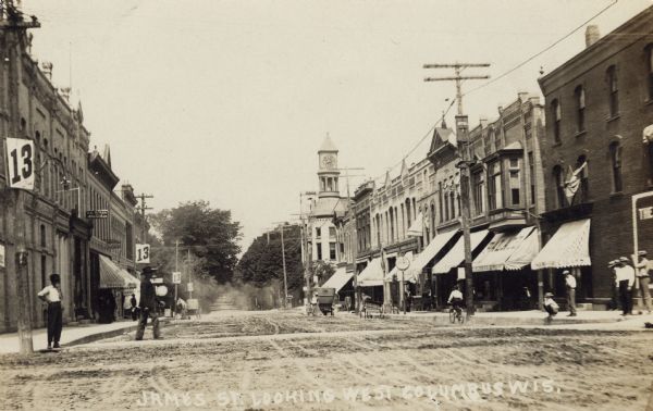 Black and white photograph looking west along James Street, with pedestrians, a horse-drawn carriage, and an automobile. City Hall is in the right background. Caption reads: "James St. Looking West, Columbus Wis."