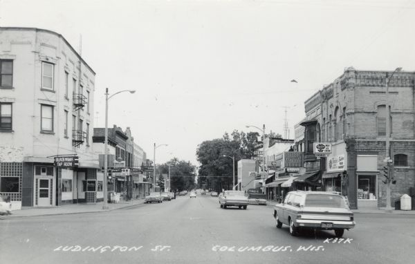 Photographic postcard of Ludington Street at an intersection, with automobiles and local businesses. Caption reads: "Ludington St., Columbus, Wis."
