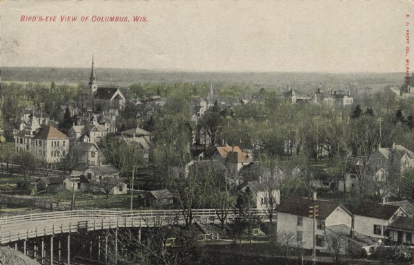 Color-enhanced black and white photographic elevated view looking across a bridge over the railroad towards the town. Caption reads: "Bird's-Eye View of Columbus, Wis."
