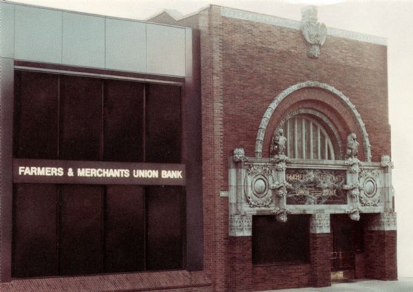 Color photographic postcard of the Farmers' and Merchants' Union Bank. The original building was designed by Louis H. Sullivan and constructed in 1919 of Crawfordsville brick and glazed terra-cotta. Additions were completed in 1960 and 1980.