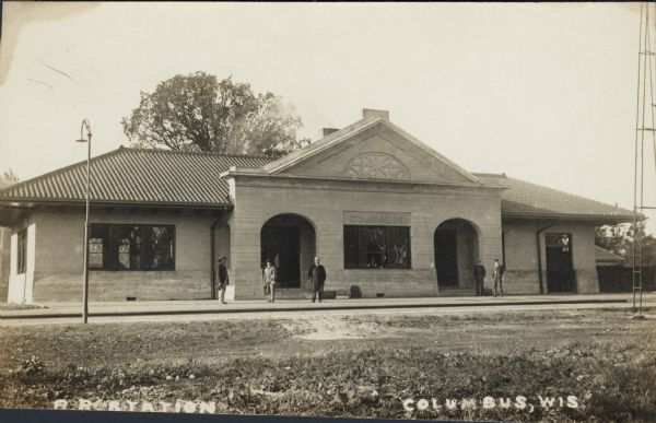 Black and white photographic postcard view across railroad tracks towards the railroad station. Five men are standing on the platform. Caption reads: "R.R. Station, Columbus, Wis."