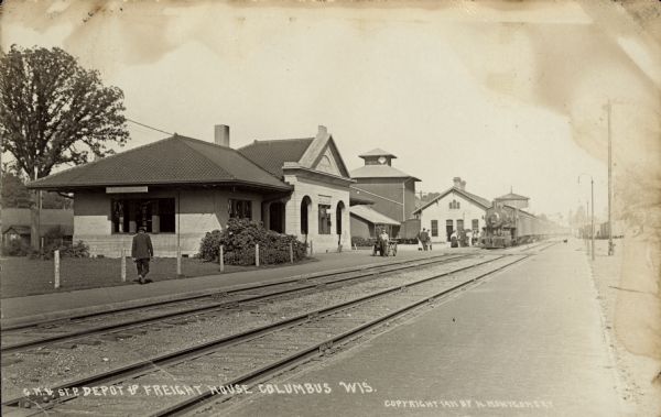 Black and white photographic postcard of a view looking across the railroad tracks at the Chicago, Milwaukee, and St. Paul depot and freight house, with a train on the tracks. Caption reads: "C. M. & St. P. Dept & Freight House, Columbus, Wis."