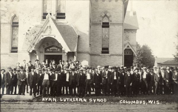 Black and white photographic postcard of men gathered in front of the church. Caption reads: "Evan. Lutheran Synod, Columbus, Wis."