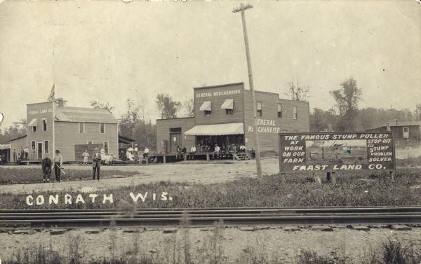 View across the railroad tracks towards downtown area, including The Faast Land Company and General Merchandise store, as well as a sign for the Faast Land Company stump puller. Several people are posing along the road and on the porches of buildings. A billboard on the right reads: "The Famous Stump Puller at Work on our Farm. Stop off see Stump Problem Solved. Faast Land Co." Caption reads: "Conrath, Wis."