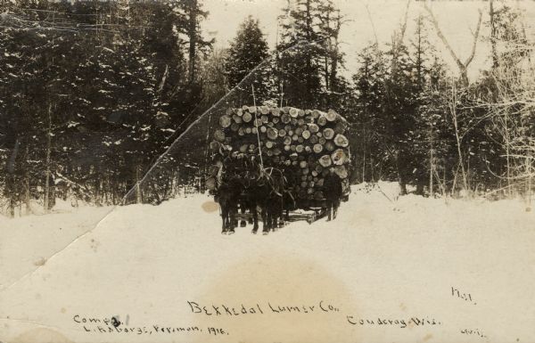 Black and white photographic postcard of a horse-drawn sled loaded with logs coming out of the woods. One or two men are driving the sled, and a man is walking next to the sled. Caption reads: "Bekkedal Lumber Company, Couderay, Wisconsin.