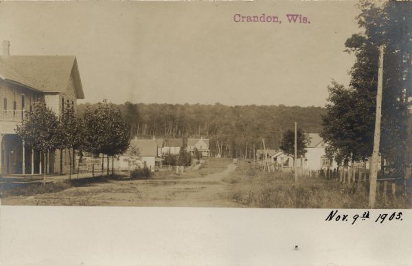 View looking up unpaved street and sidewalk of town, with dwellings on both sides, and a forested hill in the background. Caption reads: "Crandon, Wis."