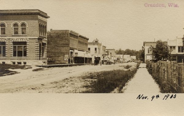 Black & white photographic postcard looking up unpaved street towards commercial buildings along the sidewalk of Main Street. Caption reads: "Crandon, Wis."
