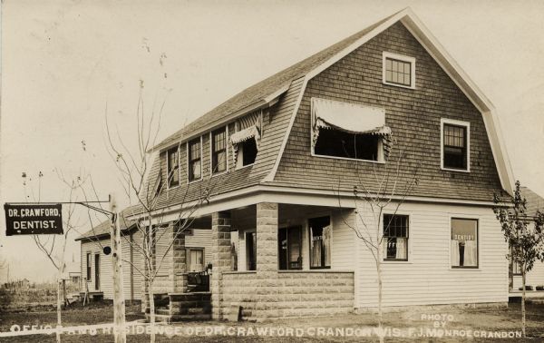 Photographic postcard showing the dentist office and residence of Dr. Crawford. There is a sign on a post in front for Dr. Crawford, Dentist, and signs on three of the windows also read: "Dentist." There is a small wood frame building in the background. Caption reads: "Office and Residence of Dr. Crawford, Crandon, Wis."