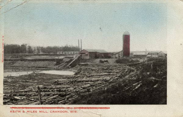 Colorized photographic postcard showing Keith & Hiles lumber mill. There is a tall building on the right that appears to be a silo. Caption reads: "Keith & Hiles Mill, Crandon, Wis."