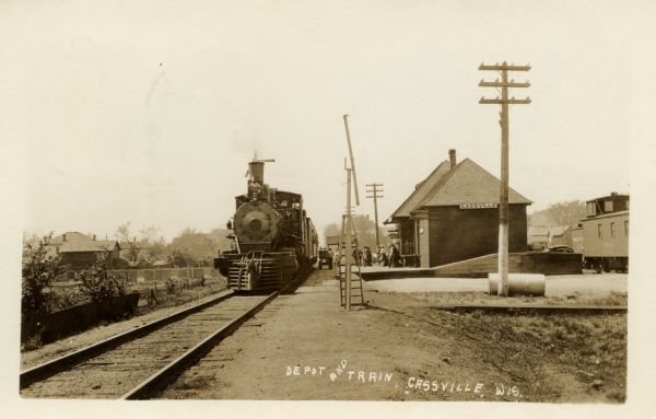 Photographic postcard of a locomotive at the head of a passenger train at the depot. Caption reads: "Depot and Train, Cassville, Wis."