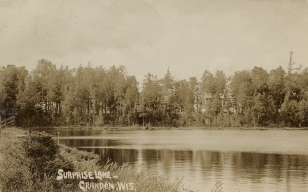 Black & white photographic postcard showing wooded shoreline of Surprise Lake. There is a two-story house behind trees on the opposite shoreline. Caption reads: "Surprise Lake, Crandon, Wis."