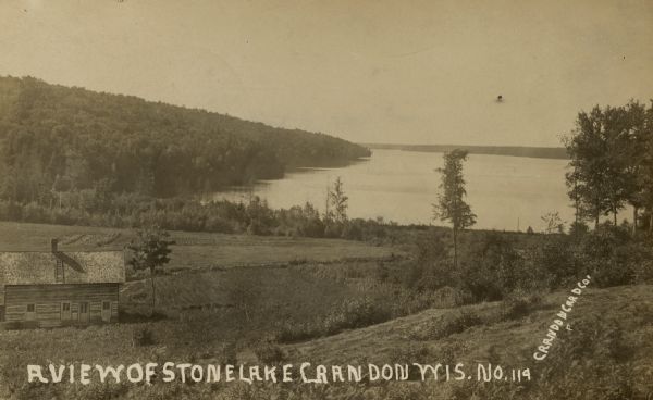 View looking down hill towards Stone Lake, with wooded shoreline and a two-story log building in the foreground. Caption reads: "A View of Stone Lake, Crandon, Wis."