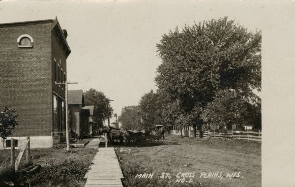 View down Main Street, a dirt road with horses and buggies and a wooden walkway. There is a brick building in the left foreground, with houses further down the walkway. There are fences and trees on the right side.