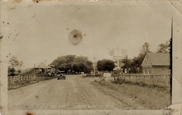Photographic postcard view looking down center of dirt highway, with a car approaching. There is a gas station on the right just past a sign reading "Cross Plains."