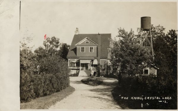 Photograph postcard view down road toward a house and water tower surrounded by shrubs and trees. Caption reads: "'The Knoll' Crystal Lake, Westfield, Wis."