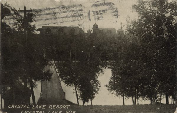 Photographic postcard view from hill through trees and over a bridge toward the Crystal Lake Resort on the opposite shoreline. There are two women wearing white dresses walking on the bridge. Caption reads: "Crystal Lake Resort, Crystal Lake, Wis." 