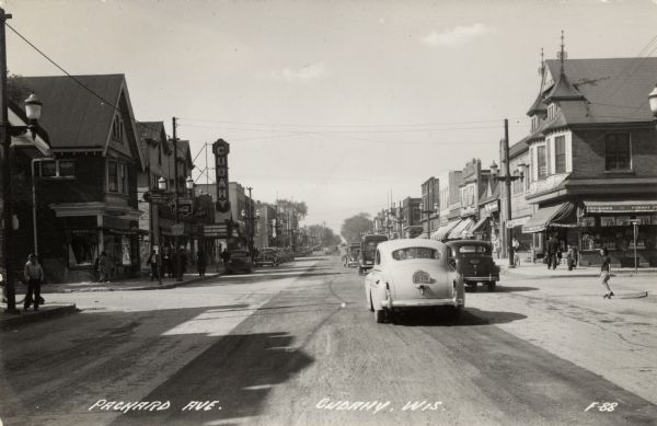 Photographic postcard view looking north on Barnard Avenue from Packard Avenue. There are businesses, automobiles and pedestrians on both sides of the street. Caption reads: "Packard Avenue, Cudahy, Wis."