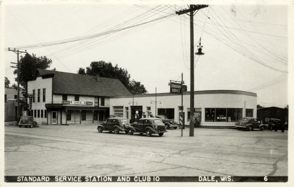 Photographic postcard view from street of the Standard Service Station, with cars parked along the curb, and another car at a pump. The building to the left is Club 10. Caption reads: "Standard Service Station and Club 10, Dale, Wis."