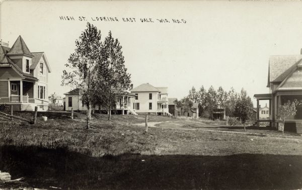 Photographic postcard of wooden, two-story houses on curved road. Two small trees are in front of the house on the left. There is a grassy area in the foreground. Caption reads: "High St. Looking East, Dale, Wis."