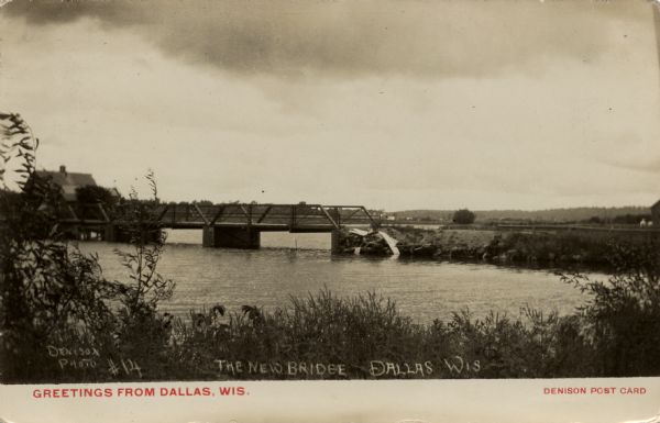 Photographic postcard view from shoreline towards a pony truss bridge over a body of water. Caption reads: "Greetings from Dallas, Wis." Written on the photograph: The New Bridge, Dallas, Wis."