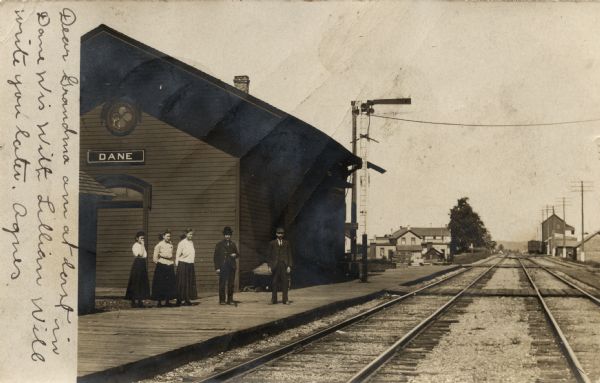 View across railroad tracks towards the train station of Dane, with three women and two men standing on the platform. Two sets of tracks stretch to the horizon. There are houses and buildings in the background.  