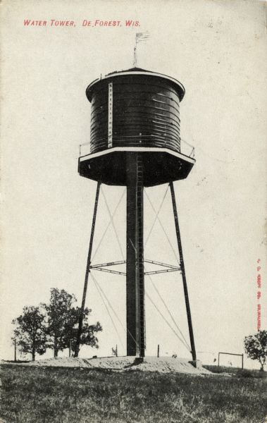 View up hill towards a water tower with a flag flying from a pole at the top. Caption reads: "Water Tower, De Forest, Wis."