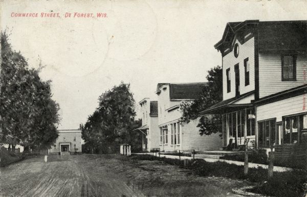 View down center of unpaved Commerce Street with buildings along a sidewalk on the right, and a building in the distance. Caption reads: "Commerce Street, De Forest, Wis."