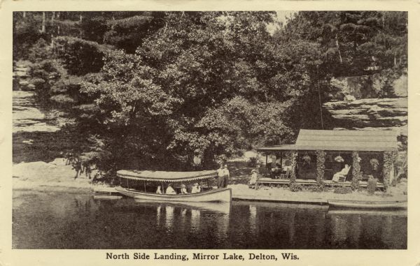 View across water towards the North Side Landing. There is a covered tour boat at the edge of the lake and passengers on board. At the shoreline is a cabin with people sitting on rocking chairs on the porch. Vines cover the cabin's posts, and planters are hanging from the ceiling. On the left side of the cabin is a dirt road between rock formations that rise above the lake. Caption reads: "North Side Landing, Mirror Lake, Delton, Wis."