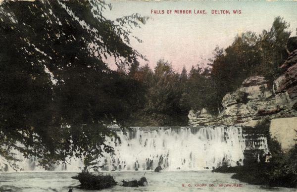 View across water towards the dam or waterfall across Mirror Lake. There are tree branches in the foreground on the left, and rock formations above the falls on the right. Caption reads: "Falls of Mirror Lake, Delton, Wis."