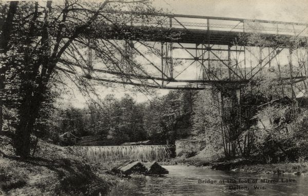 View from shoreline looking up at the bridge, and the dam. There is a building at the base on the bridge on the opposite shoreline on the right, and another building in the background. Caption reads: "Bridge at the Foot of Mirror Lake, Delton, Wis."
