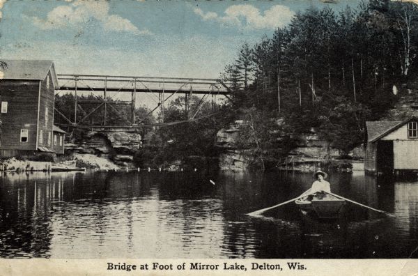View across water towards wooden buildings on the left and right, and a bridge over the foot of Mirror Lake. There is a woman in a rowboat on the lake in the foreground. Trees are along the shoreline growing on the Dells layered granite rocks. Caption reads: "Bridge at Foot of Mirror Lake, Delton, Wis."