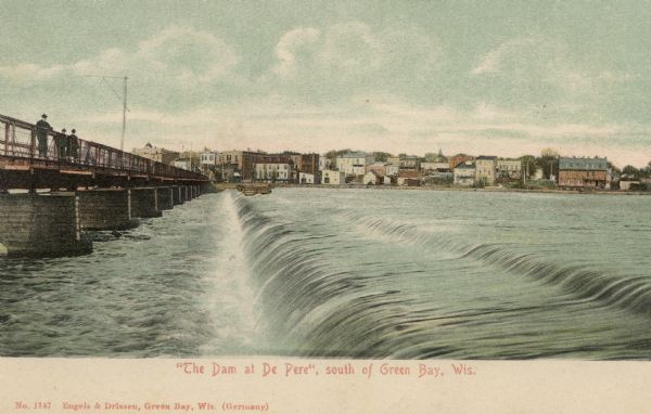 Hand-colored postcard view of Fox River dam at De Pere. Men are walking along the bridge on the left. Caption reads: "'The Dam at De Pere,' South of Green Bay, Wis."