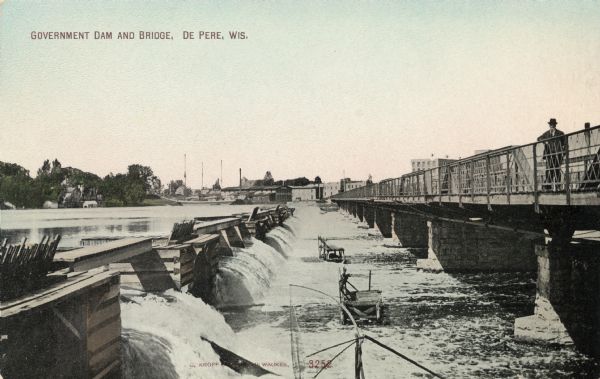 View across river, with a man standing on the bridge on the right, looking at the government dam on the left. There is a sailing ship with three masts docked on the opposite shoreline. Caption reads: "Government Dam and Bridge, De Pere, Wis."