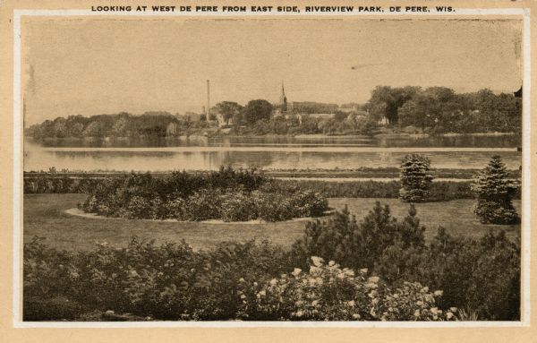 Looking at west De Pere from the east side. Shrubs and foliage are in the foreground. There are buildings and trees on the opposite shoreline. Caption reads: "Looking at West De Pere from East Side, Rivervew Park, De Pere, Wis."