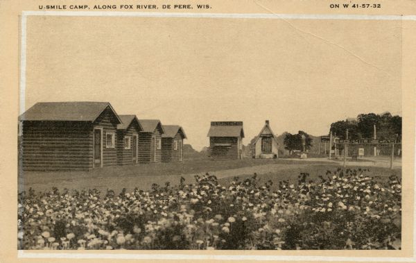 View from field of flowers towards U. Smile Camp, with four log cabins in a row on the left, another in the background, and another building next to it with a tall roof. There is a road on the right. Caption reads: "U-Smile Camp, Along Fox River, De Pere, Wis." "On W 41-57-32."