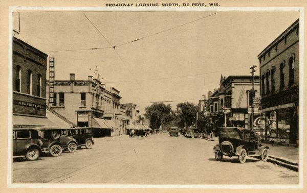 View down center of Broadway, looking north, in the business district. Automobiles are parked at an angle near the curbs, above overhead streetcar wires. Caption reads: "Broadway, Looking North, De Pere, Wis."