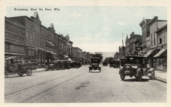 View down center of street towards automobiles and trucks parked at an angle along the curbs in the central business district. Caption reads: "Broadway, East De Pere, Wis."
