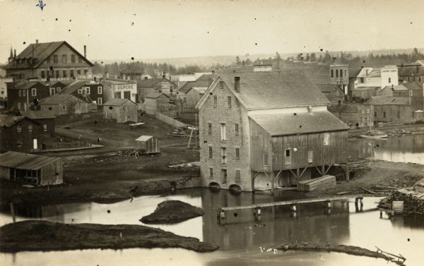 Photographic postcard of elevated view of the Wilcox Stone Mill, and part of Broadway showing the rear view of B.F. Smith's store below the hand-drawn arrow at top left. "Decades of the disposal of waste from mills and iron furnaces spanning the Fox River, created many small islands.