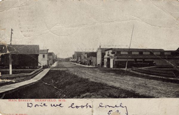 View down Main Street from between wooden sidewalk on the left, and the unpaved street on the right. Further down are commercial buildings lining both sides of the street. Caption reads: "Main Street, Deerfield, Wis."