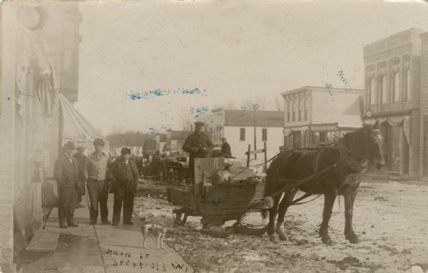 View down sidewalk along unpaved, snow-covered Main Street. Men are standing on the sidewalk, and near them is a man standing on a horse-drawn delivery sleigh. There is a dog standing on the sidewalk in the foreground. Caption reads: "Main Street, Deerfield, Wis."