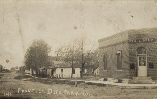 Black and white photographic postcard of unpaved Front Street, with the State Bank building on the corner on the right. Caption reads: "Front St., Deer Park, Wis."