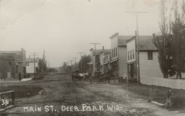 Black and white photographic postcard looking down the center of unpaved Main Street. People are standing in front of commercial buildings on the right, near horse-drawn wagons. Caption reads: "Main St., Deer Park, Wis."